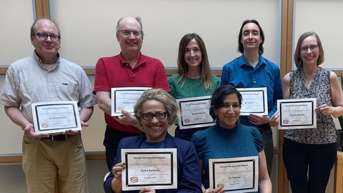 Staff honorees holding their service certificates, two seated in front and 5 standing behind them