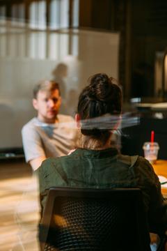 Two people seated in conversation by a table in front of a blank whiteboard