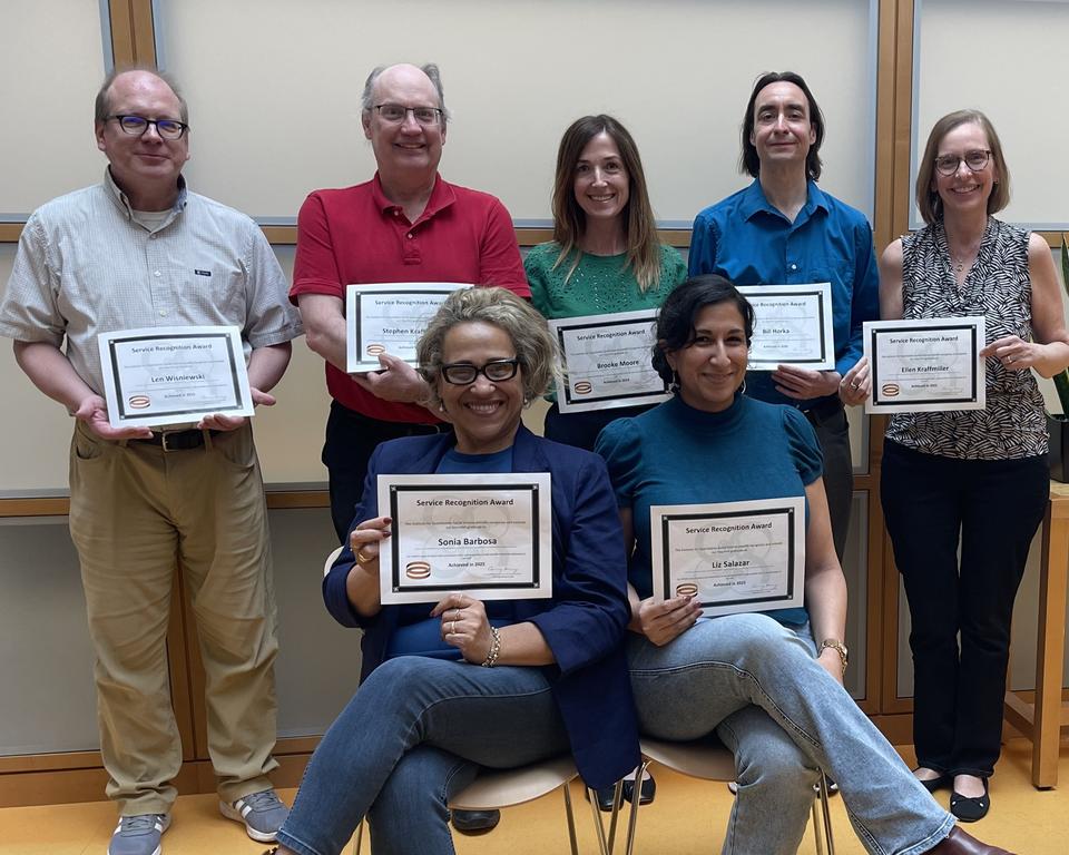 Staff honorees holding their service certificates, two seated in front and 5 standing behind them